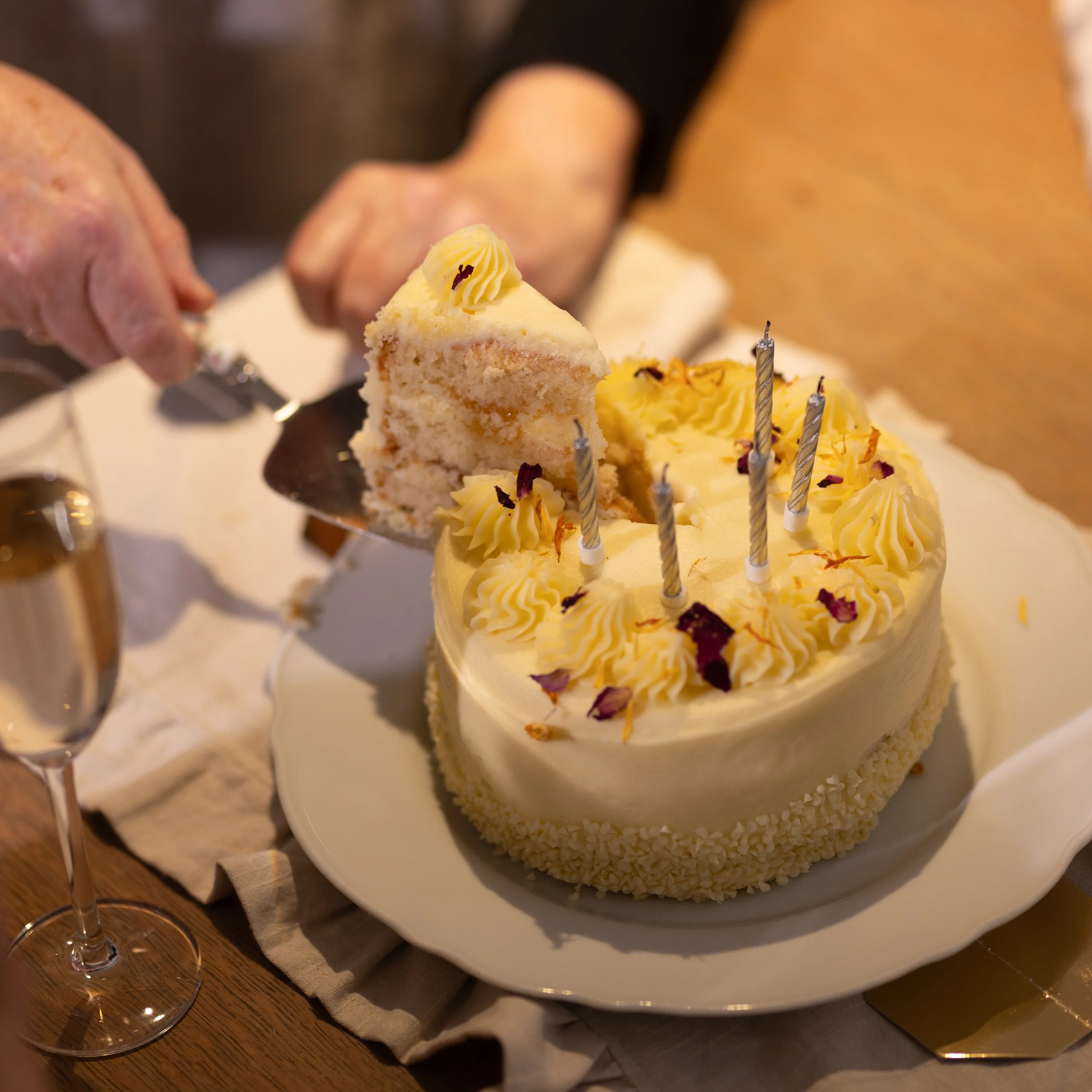 A person slices a white iced cake with yellow decorations and several lit candles on a white plate, next to a glass of champagne on a wooden table.
