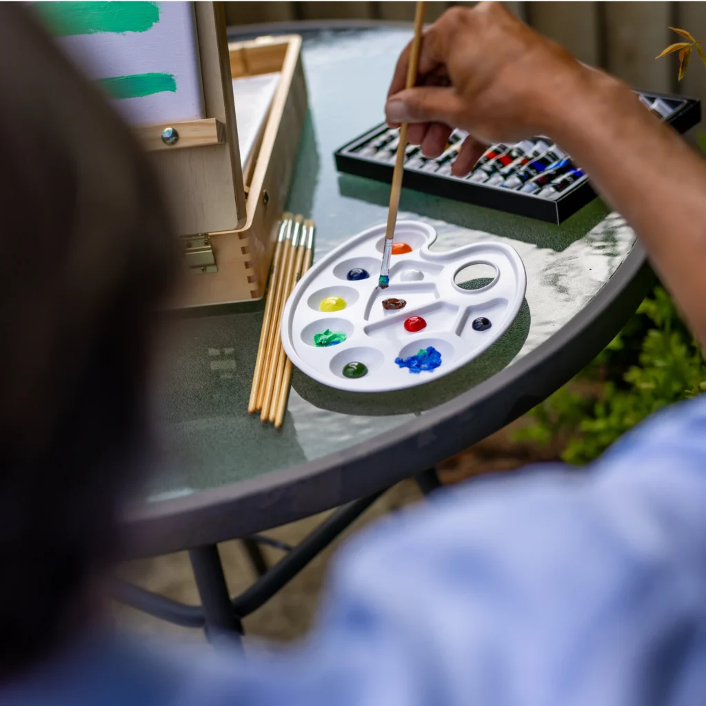 A person dips a paintbrush into a palette with various paint colours on a round outdoor table, with art supplies and a painting setup visible.
