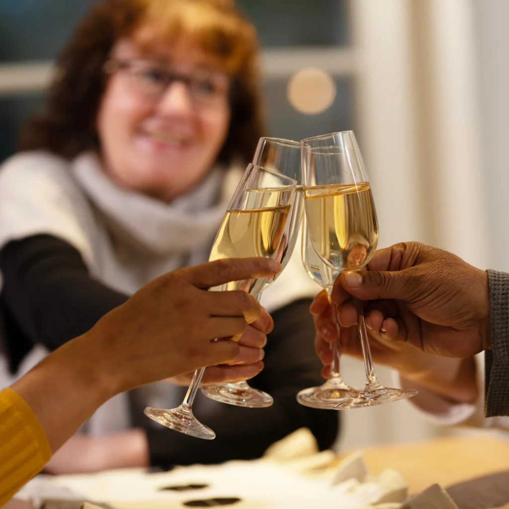 Three people clink champagne glasses together in a toast, with a smiling woman in the background. The scene suggests a celebratory or festive occasion.