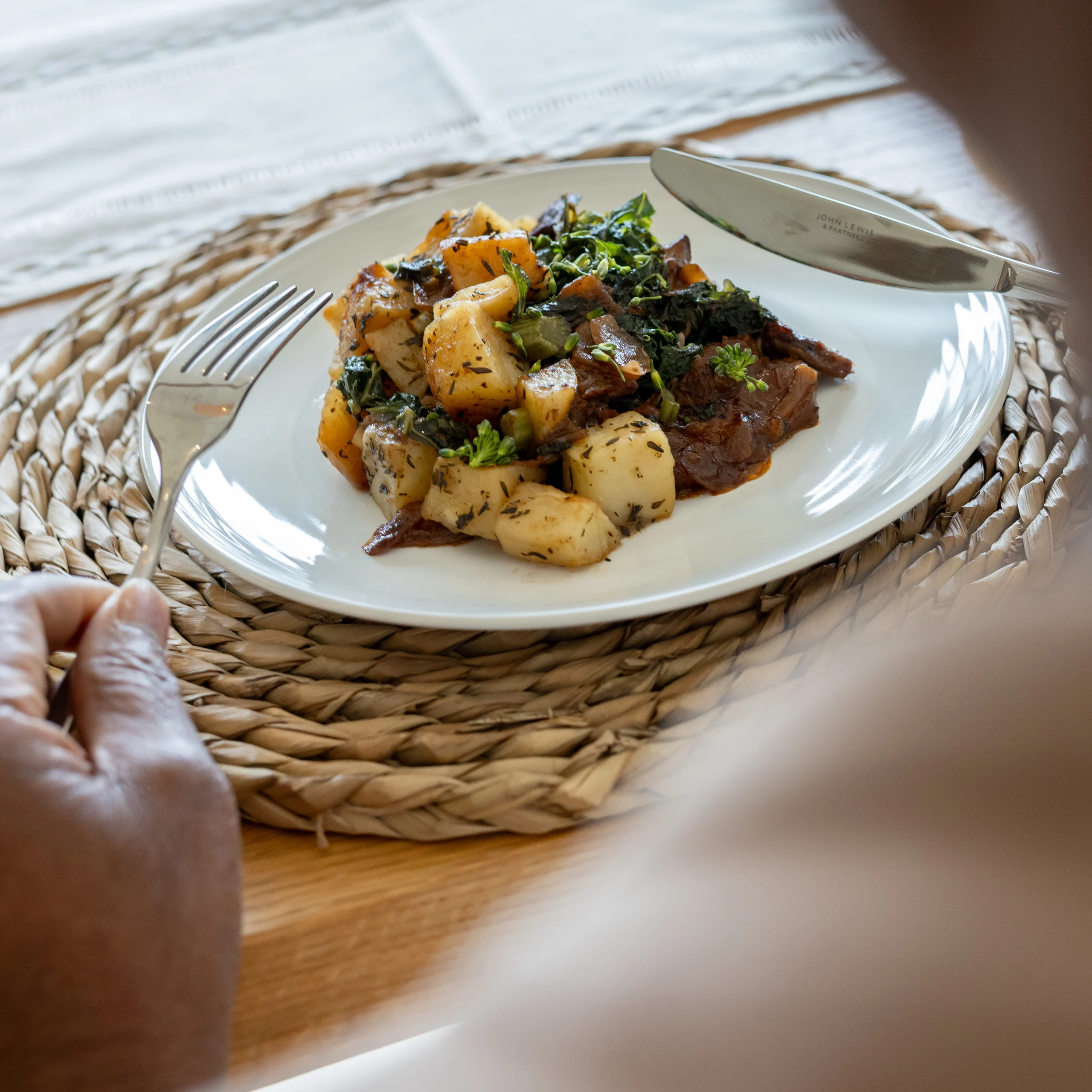A person holding a fork and knife sits at a table with a white plate of roast potatoes, vegetables, and greens on a woven mat.