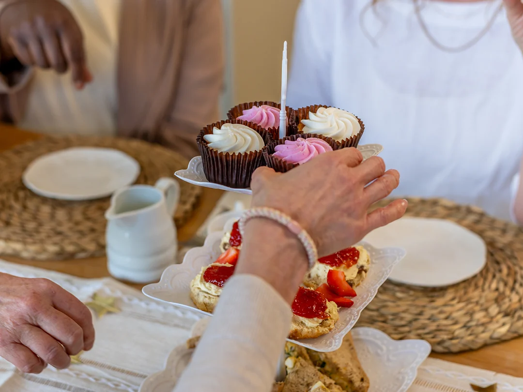 A person serves cupcakes with pink and white icing from a tiered stand at a table set with woven placemats, plates, and scones topped with cream and strawberries. Two other people are seated nearby.