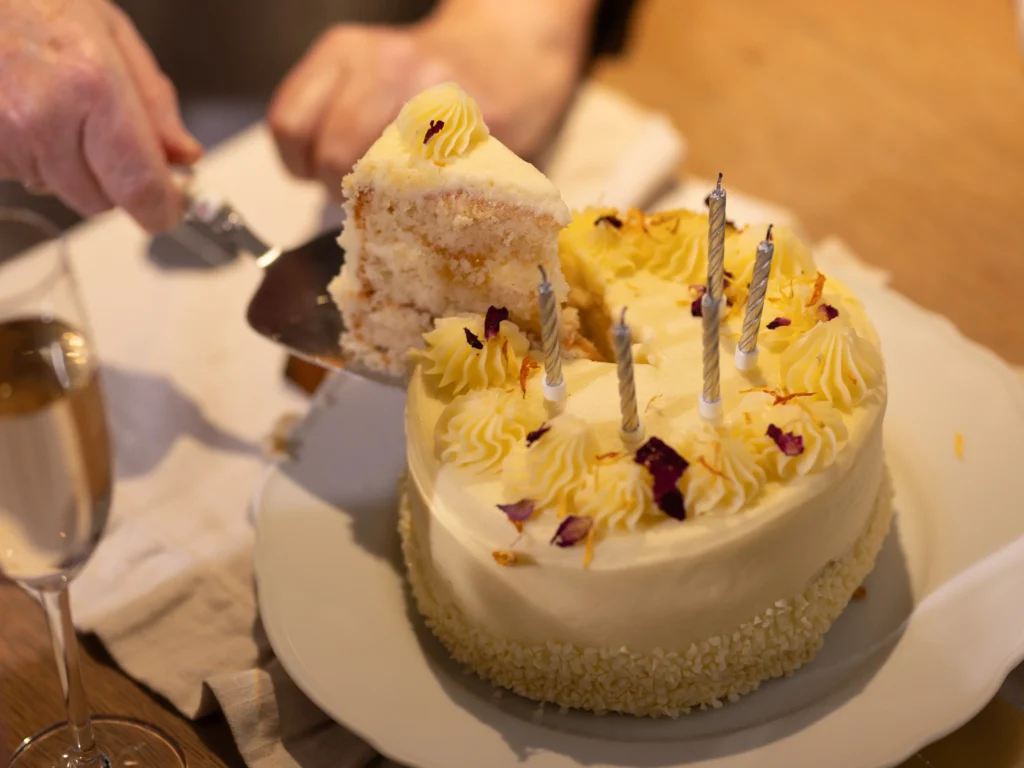 A hand lifts a slice of white-iced cake with yellow icing and four unlit candles from a cake on a white plate. A glass of champagne sits nearby on a wooden table.