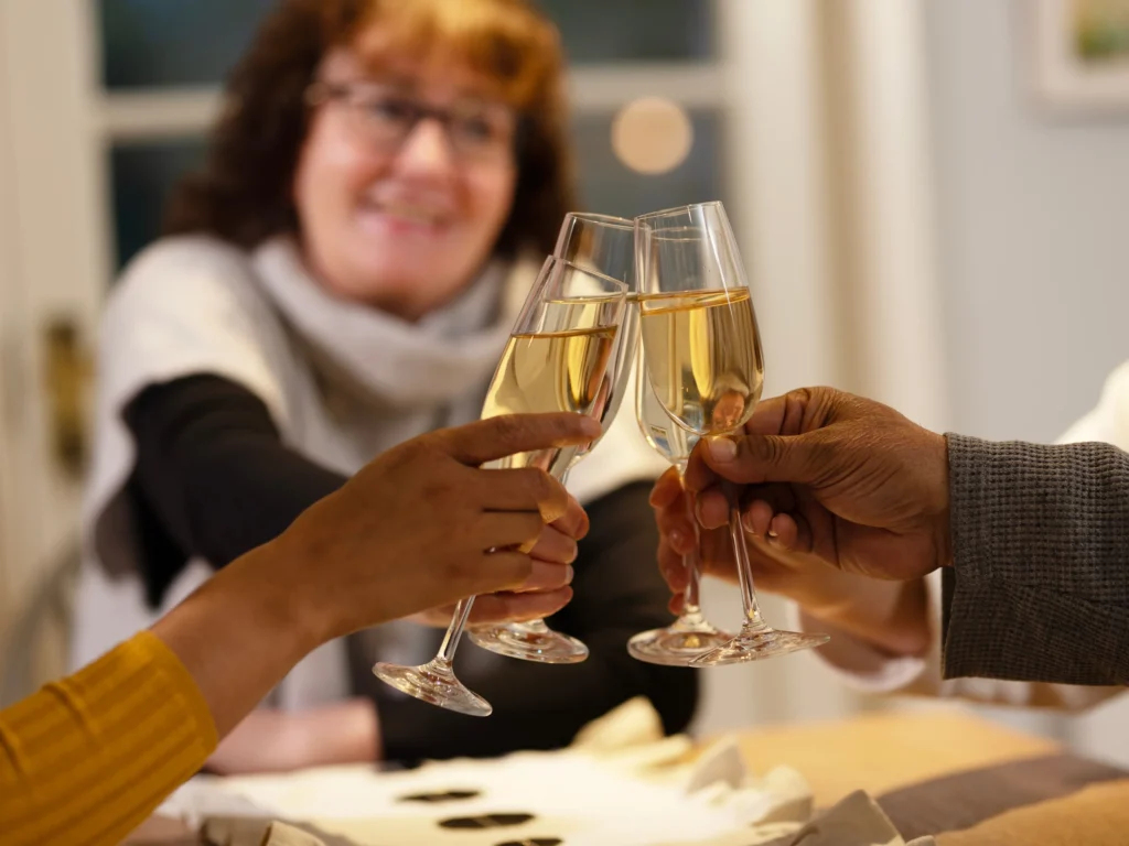 Three hands clink champagne glasses in a toast, with a smiling woman wearing spectacles and a scarf sitting in the background—capturing the warmth of Christmas activities for the elderly at home.