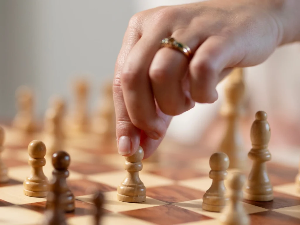 A close-up of a hand wearing a ring, moving a white pawn on a wooden chessboard. Other chess pieces are visible on the board, with soft lighting in the background—symbolising how carers can support New Year’s resolutions through thoughtful moves.