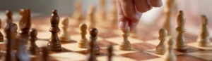A close-up of a hand moving a white pawn on a wooden chessboard, with chess pieces arranged for a game.