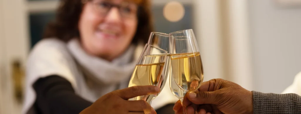 Three hands clink glasses of white wine or champagne in a toast, with a smiling woman in the background, slightly out of focus—celebrating how carers can support New Year’s resolutions together.