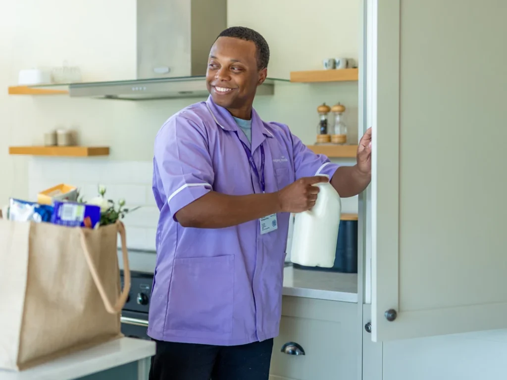 A man in a purple uniform smiles whilst placing a bottle of milk into a fridge. A brown shopping bag filled with groceries is on the worktop in a modern kitchen.