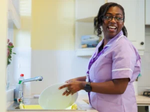A woman in a light purple uniform and glasses smiles while washing dishes at a kitchen sink, holding a plate above a draining rack.
