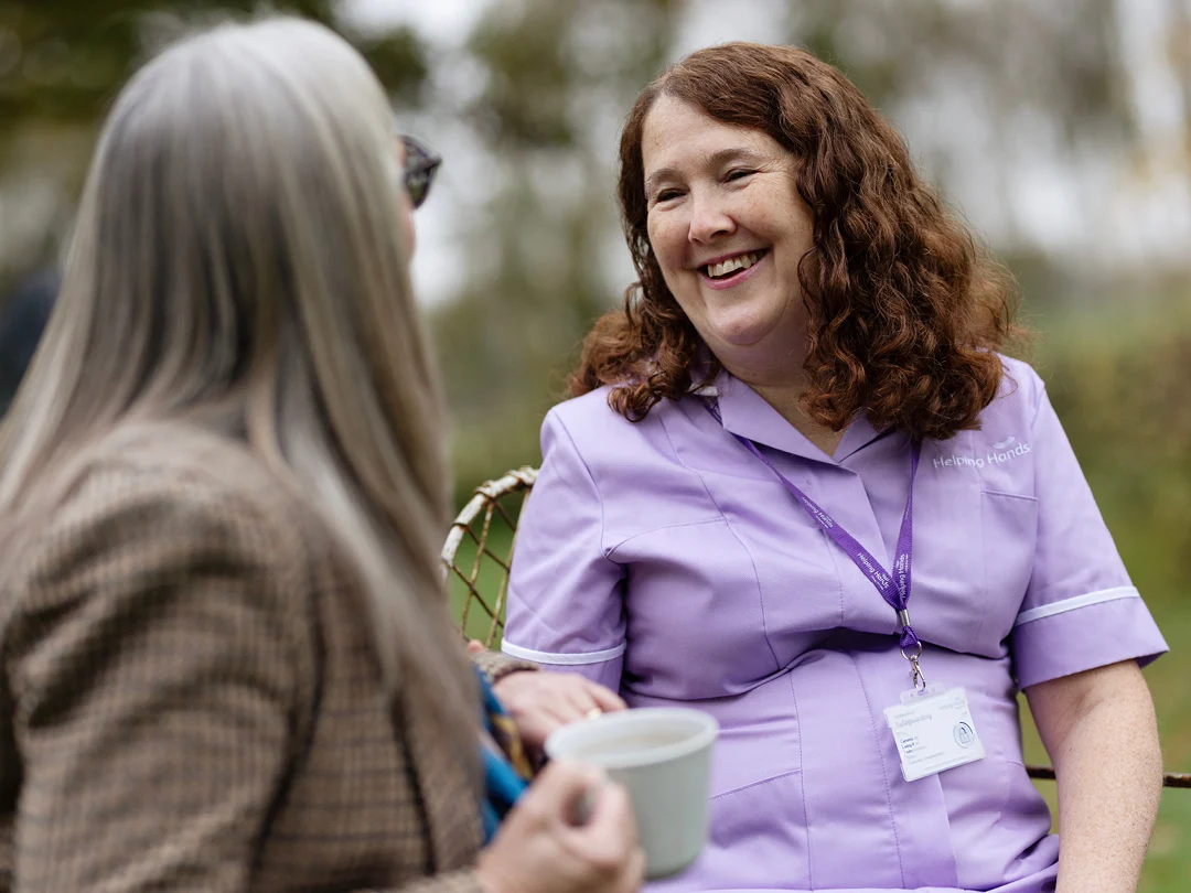 A woman in a lavender uniform with a lanyard and ID badge smiles warmly at another woman holding a cup, as they sit and talk outdoors.