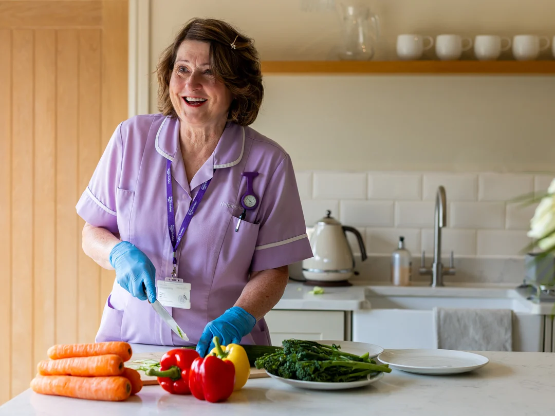 An older woman in a lavender uniform and blue gloves smiles whilst preparing vegetables, including carrots, peppers, and greens, in a bright kitchen. A kettle and cups are visible in the background.