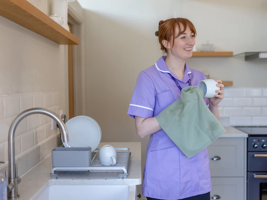 A woman in a light purple uniform stands in a kitchen, smiling while drying a white mug with a green tea towel. Clean crockery is in a draining rack beside her on the worktop.