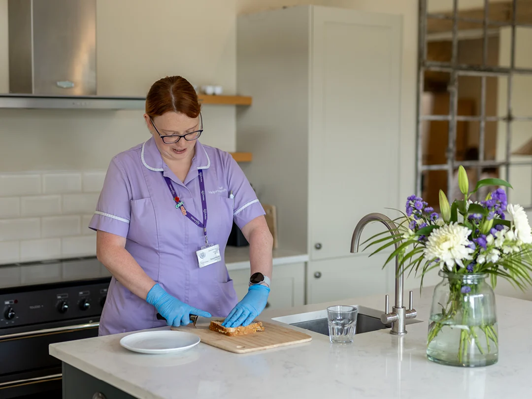 A woman in a light purple uniform and blue gloves prepares food on a chopping board in a modern kitchen. A vase of white and purple flowers is on the worktop beside her.