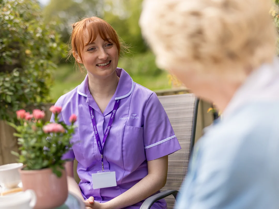 A young woman in a purple nurse uniform smiles whilst talking to an older woman outdoors. A potted pink flower and a teacup are on the table in front of them.