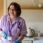 A smiling woman in a purple uniform and blue gloves stands in a kitchen, holding a cleaning cloth. She wears a name badge and a stethoscope, with shelves, a kettle, and a sink visible in the background.