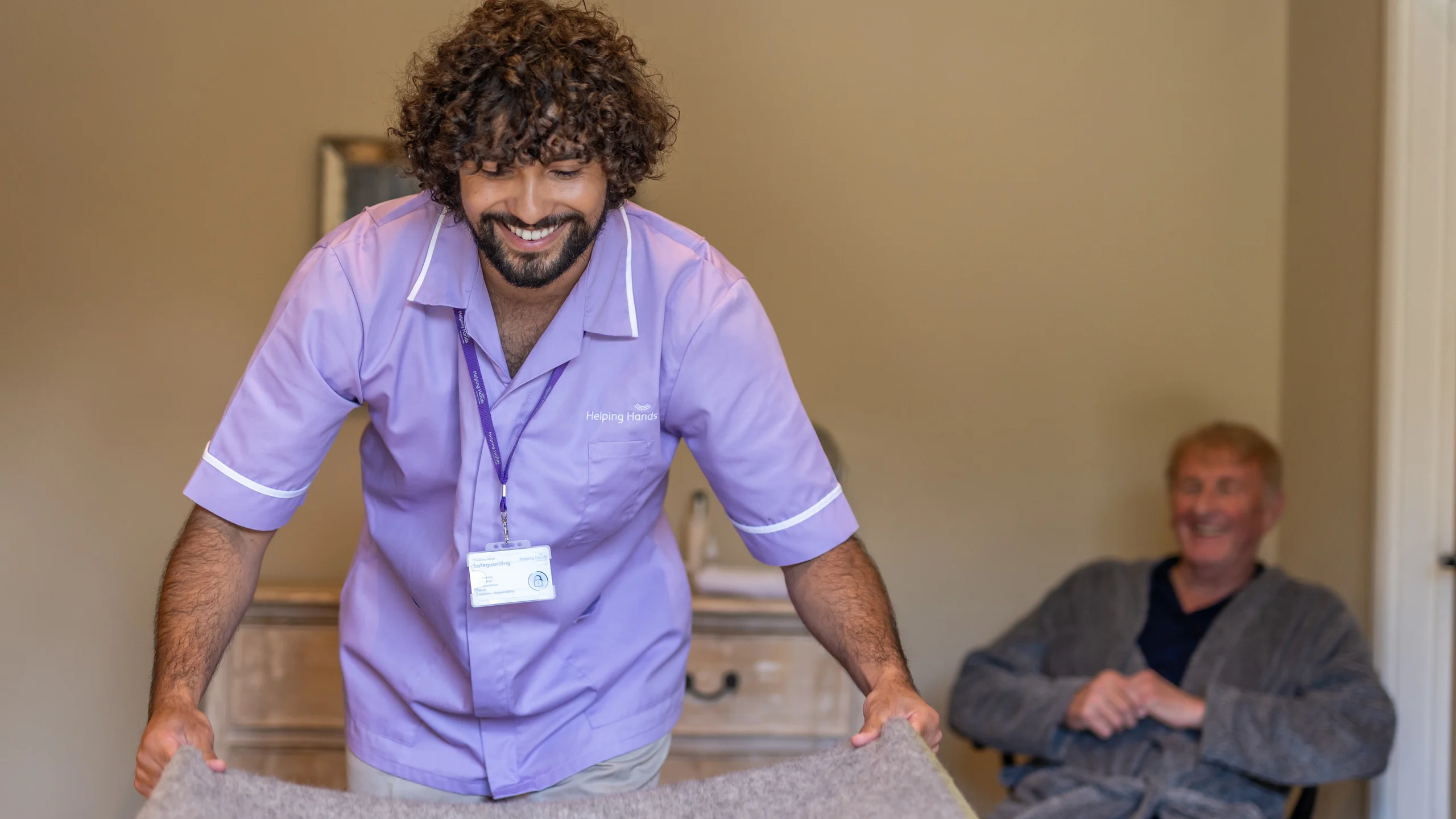 A smiling carer in a purple uniform makes a bed while an older person in a dressing gown sits and smiles in the background of a warmly lit room.