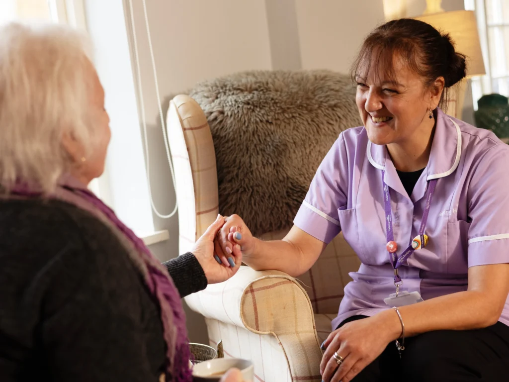 A smiling carer in a purple uniform sits holding hands with an older woman, offering comfort and support in a cosy, well-lit living room.