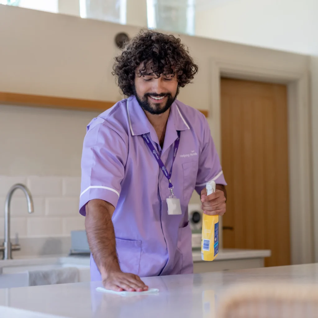 A smiling person in a light purple uniform wipes a kitchen worktop with a cloth, holding a yellow cleaning spray bottle. A sink and wooden door are visible in the background.