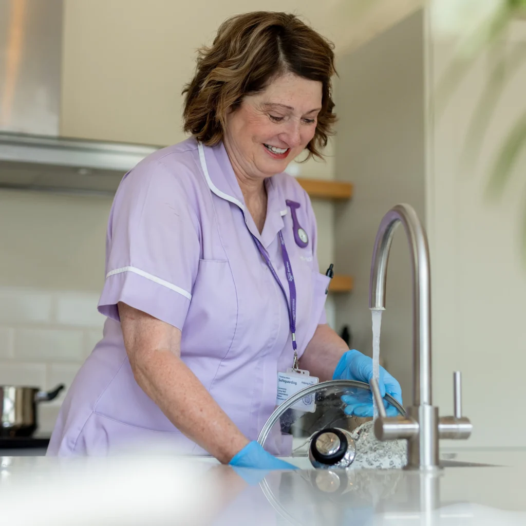 A smiling woman in a light purple uniform and blue gloves washes a glass pot lid under a running kitchen tap. She appears to be a healthcare or home care worker in a clean, modern kitchen.