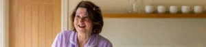 An older woman with wavy brown hair, wearing a lavender top, smiles warmly in a bright kitchen. A wooden door and a shelf with white mugs are visible in the background.