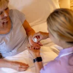 A nurse hands medication to an elderly woman sitting up in bed. The woman wears a striped shirt and smiles, while the nurse holds a pill bottle and dispenses medicine into her hand.