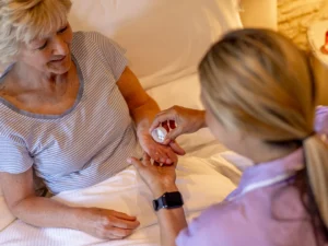 A nurse hands medication to an elderly woman sitting up in bed. The woman wears a striped shirt and smiles, while the nurse holds a pill bottle and dispenses medicine into her hand.
