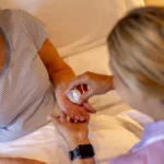 A carer helps an elderly woman in bed by giving her medication, placing tablets in her hand. The elderly woman is wearing a striped shirt and looks up while the carer assists her.