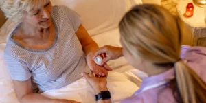 A carer helps an elderly woman in bed by giving her medication, placing tablets in her hand. The elderly woman is wearing a striped shirt and looks up while the carer assists her.