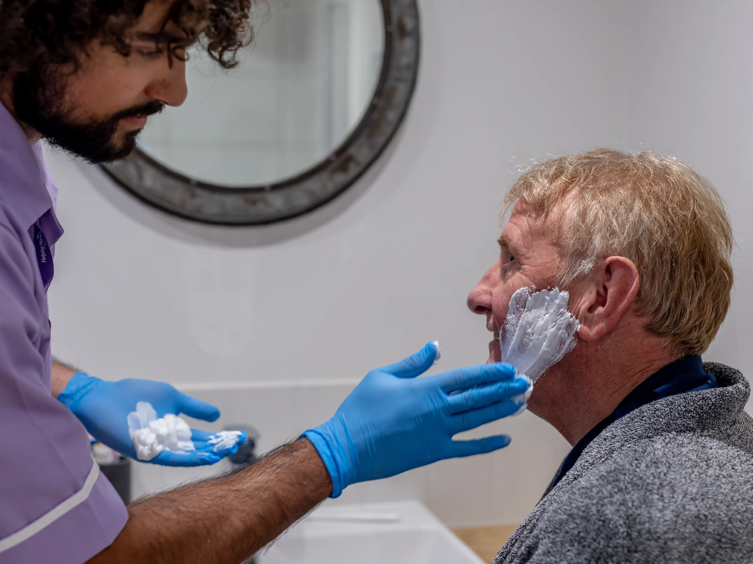 A carer wearing blue gloves applies shaving foam to an elderly man’s face as he sits by a bathroom basin, preparing for a shave. A round mirror hangs on the wall in the background.