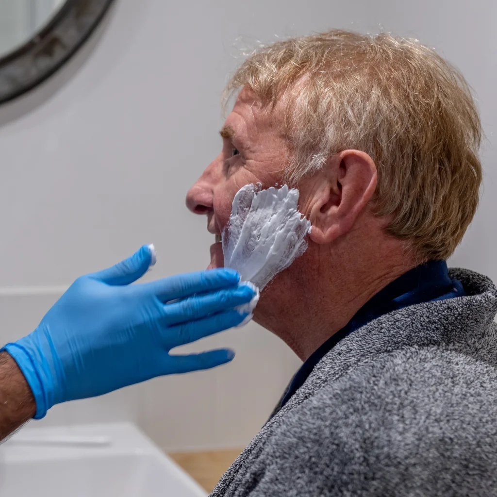 A person wearing blue gloves applies shaving foam to the cheek of an older man with fair hair, who is sitting wrapped in a grey towel, in a bathroom.