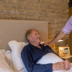 A carer in a purple uniform gently smiles and talks to an older man lying in bed, covered with a white blanket, in a cosy bedroom with a brick wall and a bedside lamp.