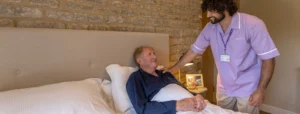 A carer in a purple uniform gently smiles and talks to an older man lying in bed, covered with a white blanket, in a cosy bedroom with a brick wall and a bedside lamp.