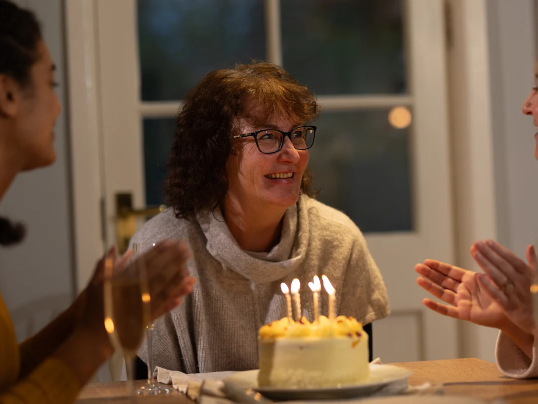 A woman with glasses smiles as she sits in front of a lit birthday cake, whilst two people beside her clap and celebrate at a table indoors.