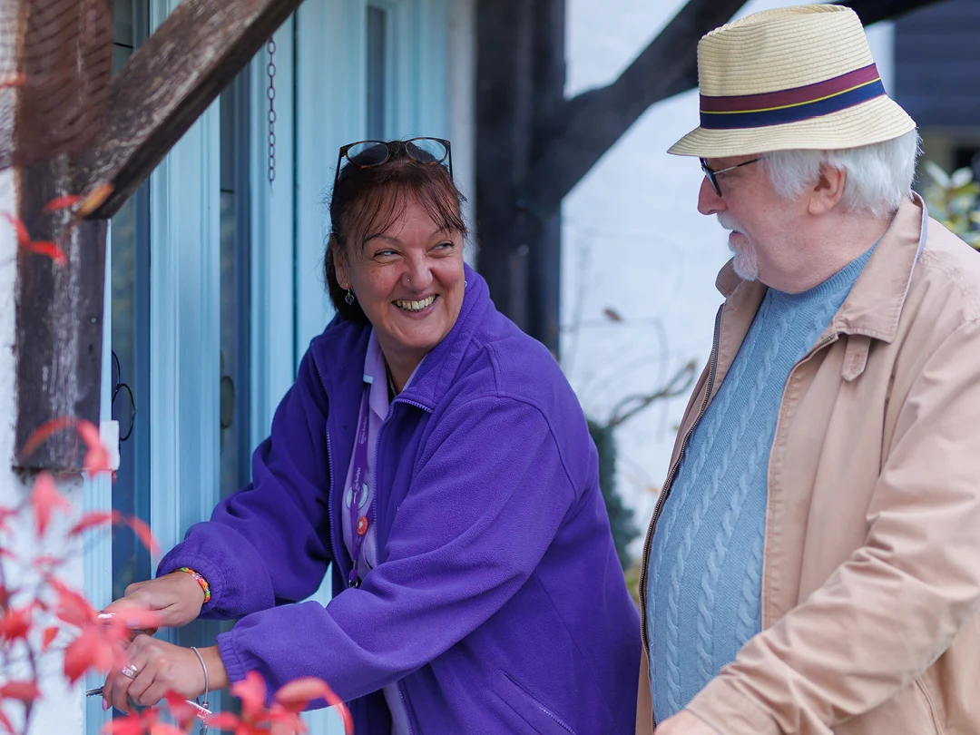 A woman in a purple jacket smiles as she unlocks a blue door, whilst an older man in a beige jacket and straw hat stands nearby, both appearing happy and engaged outside a house.