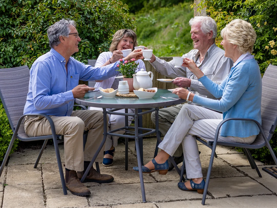 Four older adults sit around a circular outdoor table, smiling and raising teacups in a toast. Enjoying Christmas activities for the elderly at home, they share cheerful tea time together in a garden adorned with festive joy.