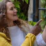 Two women, one older and one younger, smiling and pointing at something on a leafy wall outdoors. They appear happy and engaged, standing close together amid green foliage.