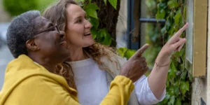 Two women, one older and one younger, smiling and pointing at something on a leafy wall outdoors. They appear happy and engaged, standing close together amid green foliage.
