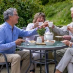 Four older adults sit around a garden table, smiling and raising teacups in a toast. The table holds a teapot, cups, and snacks, with greenery and shrubs in the background on a sunny day.