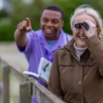 An older woman smiles while using binoculars, standing by a wooden fence outdoors. A man beside her points excitedly ahead and holds a book, suggesting they are birdwatching together in a green, natural setting.