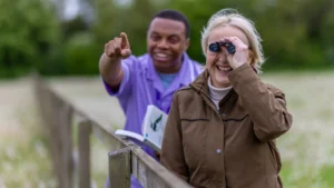 An older woman smiles while using binoculars, standing by a wooden fence outdoors. A man beside her points excitedly ahead and holds a book, suggesting they are birdwatching together in a green, natural setting.