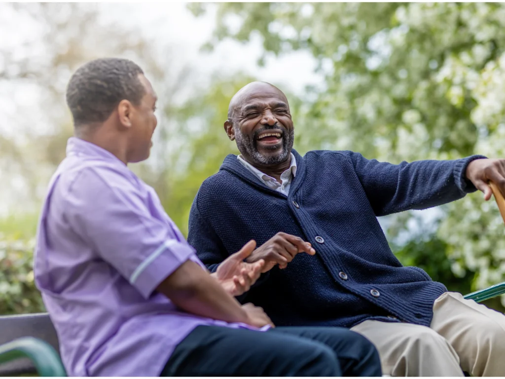 Two men sit on a bench outdoors, smiling and laughing together. One man wears a navy cardigan and holds a walking stick, while the other wears a light purple shirt. Trees with green leaves are in the background.