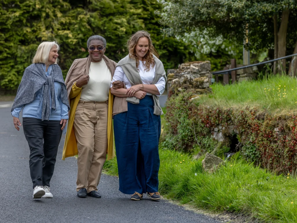 Three women of different ages walk arm-in-arm along a rural road, smiling and talking. They are dressed casually and surrounded by greenery and trees.