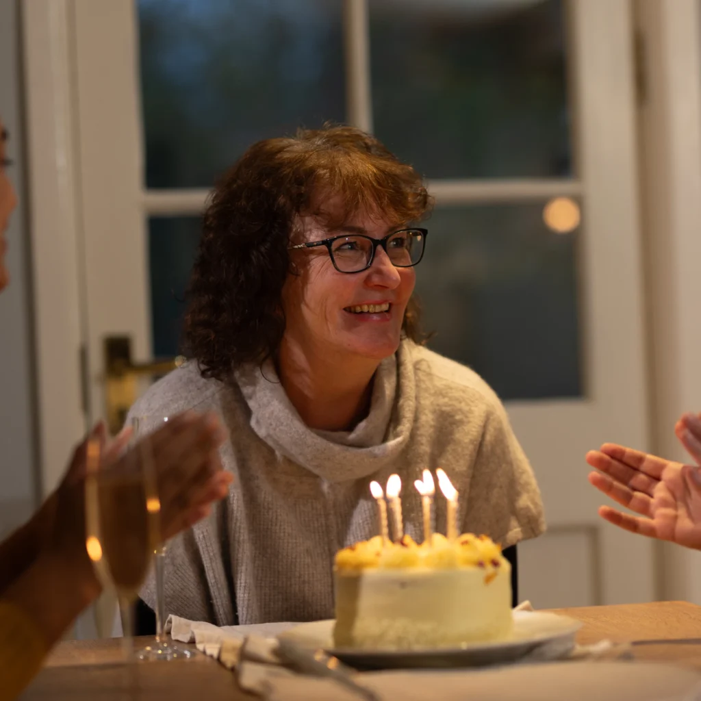 A woman with curly hair and glasses smiles at a table with a lit birthday cake in front of her, while people around her clap and celebrate in a warmly lit room.