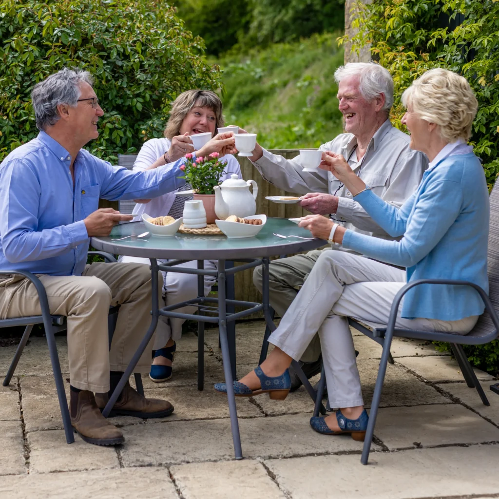 Four older adults sit outdoors around a round table, smiling and raising teacups in a toast. The table has a teapot, cups, and snacks. Greenery and sunshine create a cheerful atmosphere.