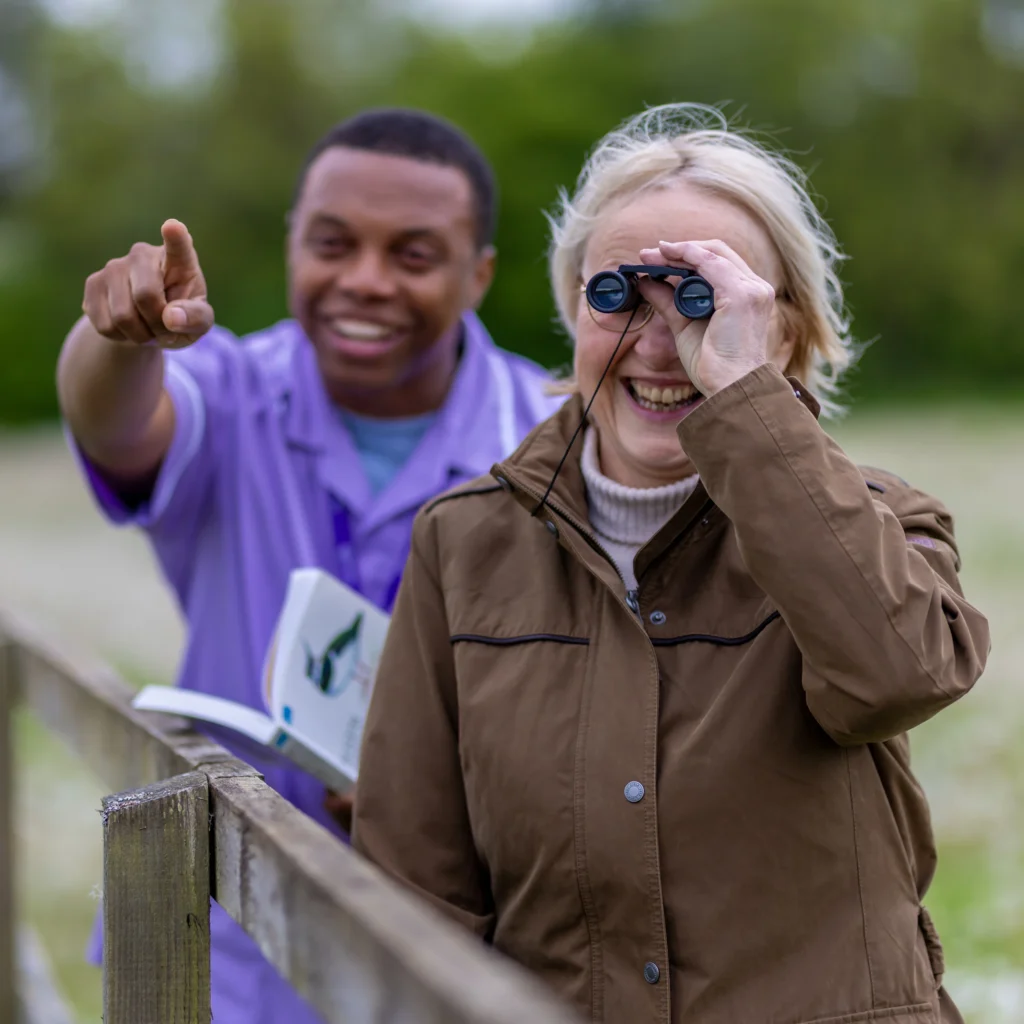 An older woman smiles while looking through binoculars. A younger man beside her points ahead and holds an open book. They are outdoors near a fence, surrounded by greenery.