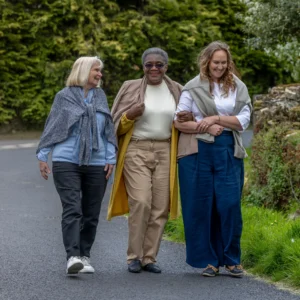 Three older women walk arm-in-arm down a paved path, smiling and enjoying each other's company, with greenery and trees in the background.