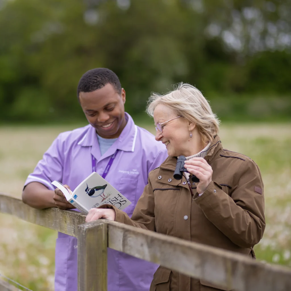 A young man in a lavender uniform and an older woman in a brown jacket stand by a wooden fence outdoors, smiling and reading a bird guidebook together. A grassy field and trees are in the background.