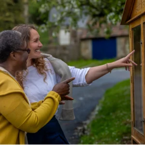 Two women smile and point at a wooden outdoor noticeboard on a path, surrounded by greenery. One woman wears a yellow jumper, while the other has a light grey jumper draped over her shoulders.