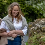 Three older women walk outdoors arm in arm, smiling and talking together. They are dressed in casual clothes and appear to be enjoying a pleasant day in a green, leafy setting.