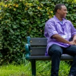 Two men sit on a park bench, smiling and talking. One holds a walking stick, and the other wears a light purple shirt with an ID badge. Green foliage fills the background, creating a relaxed outdoor setting.