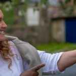 Two women smile as they read and point at notices posted on a wooden outdoor noticeboard, with greenery and a building blurred in the background.
